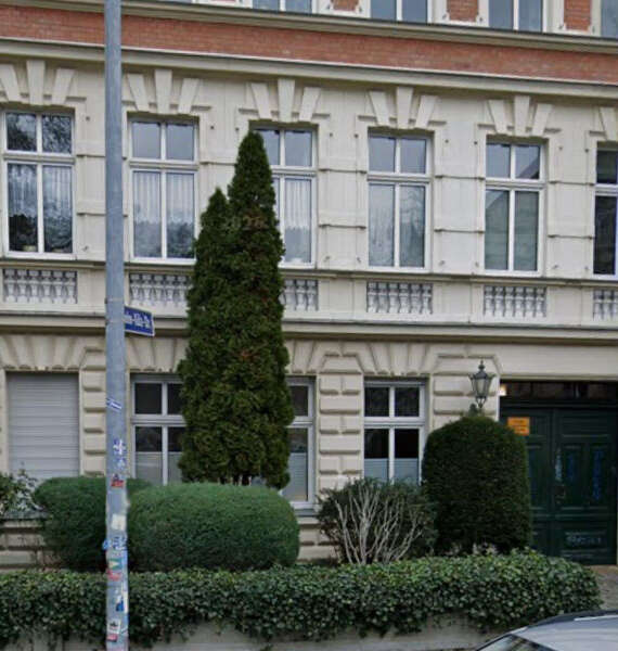 Beige, multi-story building with tall windows, a dark green door, and manicured shrubs in front; a tall evergreen tree and street sign nearby.